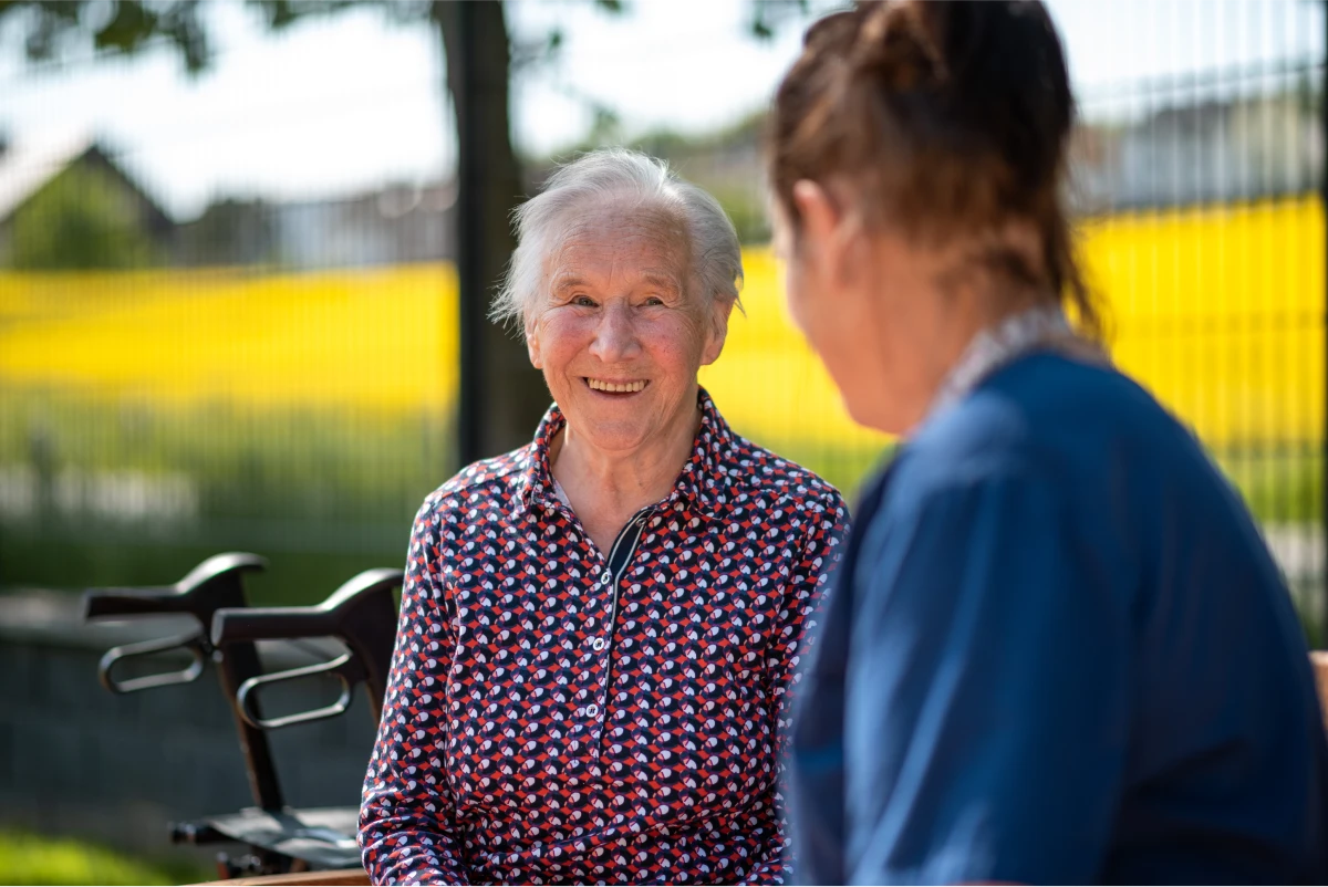 pflegedienst-altenpflege-krankenpflege-reichenbach-14 Klientin in Frontansicht draußen im Hintergrund ein verschwommenes Rapsfeld, im Vordergrund eine weibliche Person schräg von hinten angeschnitten