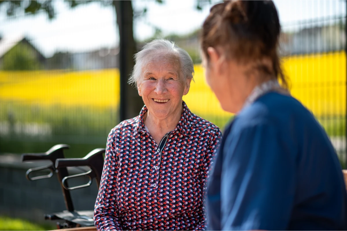 pflegedienst-altenpflege-krankenpflege-reichenbach-20 Klientin in Frontansicht draußen im Hintergrund ein verschwommenes Rapsfeld, im Vordergrund eine weibliche Person schräg von hinten angeschnitten